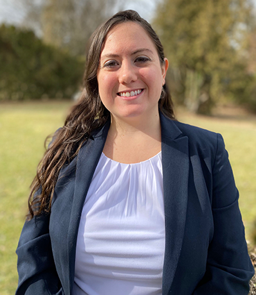 The image shows a headshot of a smiling woman, Alexandria Miller, with long, dark hair. She's wearing a dark blue blazer over a white top with a gathered neckline. The background is slightly blurred, showing a grassy outdoor setting with some trees.