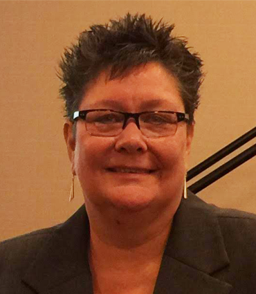 A headshot of a woman with short, dark, spiky hair and glasses. She is smiling faintly and wearing a dark suit jacket and long, slender earrings. The background is a plain, light brown wall with a dark diagonal line visible on the right side.